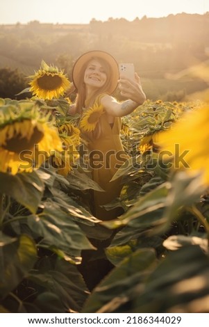 Young woman in yellow dress and straw hat taking selfie photo in blooming sunflower field at sunset. Happy girl with dimples on cheeks holding smartphone and making picture in summer flower field.