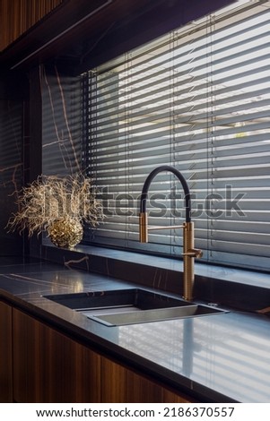 Wooden blinds black color closeup on the window. Wood slats 50mm wide. Venetian wood blinds in the kitchen. Black tapes. Sink with copper faucet near the window. Round vase is on the windowsill. Royalty-Free Stock Photo #2186370557