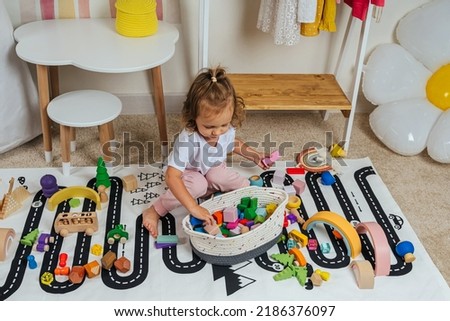 A little girl sitting on floor plays with toy wooden blocks on a rug with a picture of a road in playroom. Educational game for baby and toddler in modern nursery. 