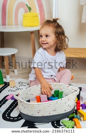 A smilling little girl sitting on floor plays with toy wooden blocks on a rug with a picture of a road in playroom. Educational game for baby and toddler in modern nursery. 