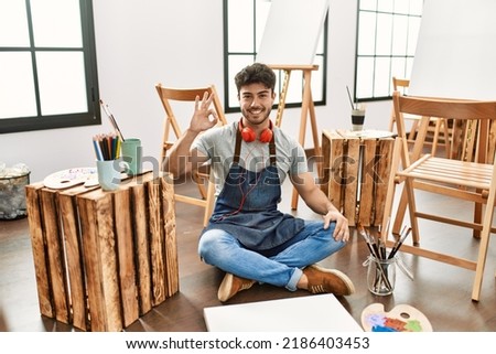 Young hispanic man sitting at art studio smiling positive doing ok sign with hand and fingers. successful expression. 