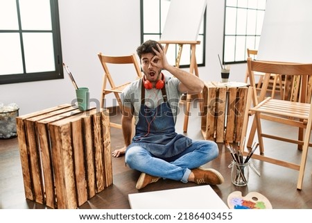 Young hispanic man sitting at art studio doing ok gesture shocked with surprised face, eye looking through fingers. unbelieving expression. 