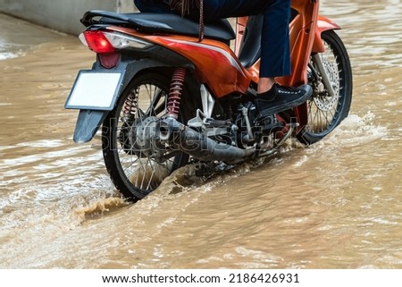 Man ride motorcycle passing through flooded road. Riding motorbike on flooded road during flood caused by torrential rains. Flooded road with large puddle. Splash by motorcycle through flood water. Royalty-Free Stock Photo #2186426931