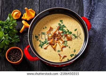 Kanttarellikeitto, Finnish Chanterelle Soup in red pot on dark wooden table with fresh Chanterelle mushrooms on background, horizontal view from above