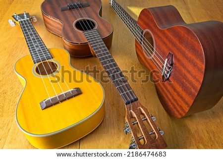 Collection of three ukuleles aligned on sun drenched wooden table top. High angle view. Side lit, showing texture of wood grains. Royalty-Free Stock Photo #2186474683