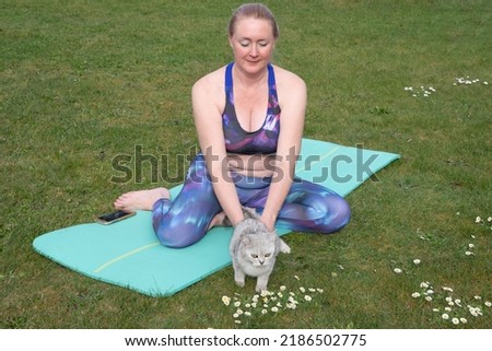 middleaged woman doing yoga with a kitten on a blue mat in the middle of a green