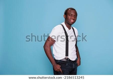 Confident man posing for camera while standing on blue background. Smiling heartily handsome young adult person with fashion style wearing white tshirt and suspenders. Studio shot
