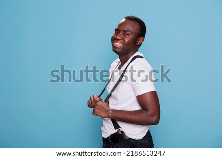 Joyful african american man smiling heartily while posing for camera on blue background. Cheerful and confident looking young boy wearting fashing clothing while pulling suspenders.