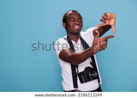 Young guy with DSLR device gesturing taking photo with hands. Confident professional photographer doing frame gesture with hands while having modern camera on blue background. Studio shot