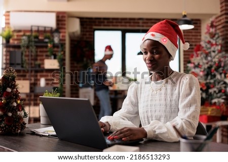 African american woman with santa hat working on business in office with christmas tree and lights. Festive employee using laptop at company job with seasonal decor and ornaments.