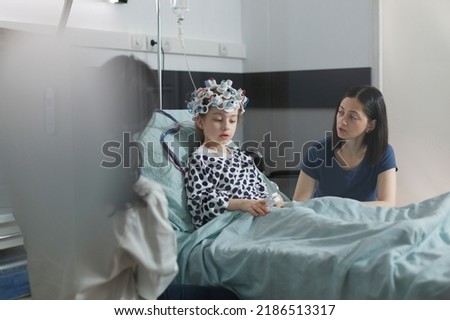 Neurology specialist examining neurological disease state of ill kid. Hospitalized sick girl wearing EEG headset resting in patient bed while doctor analyzing brain condition. Royalty-Free Stock Photo #2186513317