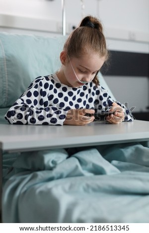 Hospitalized ill kid playing games on modern mobile phone while resting inside hospital pediatrics ward. Sick girl under treatment resting on patient bed while watching cartoons on smartphone. Royalty-Free Stock Photo #2186513345