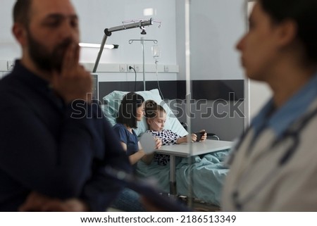 Hospitalized ill little kid watching cartoons on mobile phone while resting in hospital pediatric ward. Sick girl playing games on smartphone with mother while father talking with expert pediatrist.