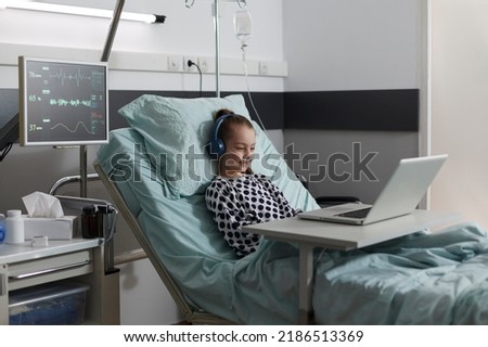 Happy sick little girl under treatment sitting on patient bed with laptop while watching cartoons inside hospital pediatrics ward room. Ill kid having oxygen tube enjoying funny videos on computer. Royalty-Free Stock Photo #2186513369