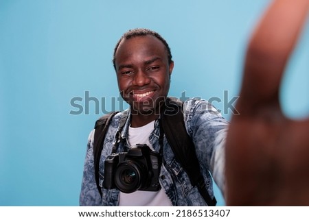 Happy photographer having camera and taking selfie while waving at camera on blue background. Confident photography enthusiast with DSLR photo device and backpack enjoying vacation.