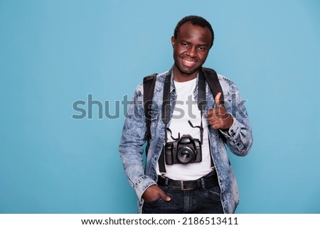 Young photographer showing thumbs up hand gesture while having professional camera on blue background. Happy photography enthusiast with DSLR photo device giving okay approve hand symbol.