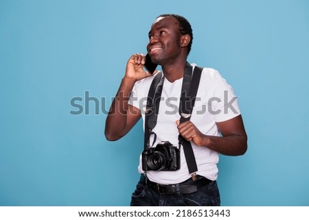 Happy professional photographer having DSLR camera talking on phone while standing and smiling on blue background. Joyful photography enthusiast talking on mobile phone while having photo device.