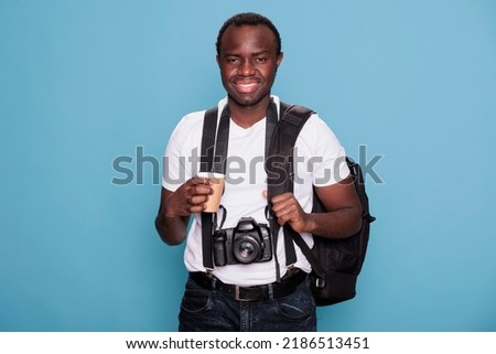 Tourist with professional camera and travel backpack ready for holiday journey on citybreak. African american photographer with DSLR photo device and travel roadtrip bag standing on blue background.