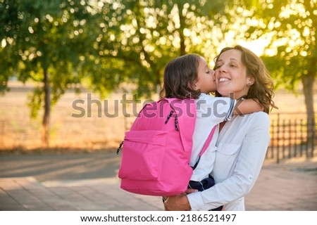 smiling mother with long brown hair and a white shirt with her daughter in her arms with a pink backpack giving her a kiss ready to start back to school