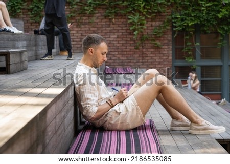 Stylish young man drawer makes sketches using pen in paper notebook. Guy finds muse in park drawing picture sitting on rug put on stylized steps. City dwellers use sunny days to rest and work