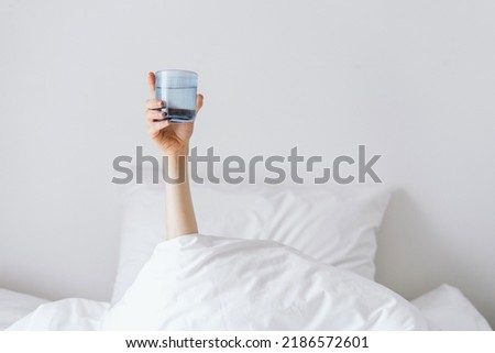 Person's hand holding glass of water when lying in bed, morning rituals, copy space, morning in contemporary hotel room, wellness and relax, purified water Royalty-Free Stock Photo #2186572601