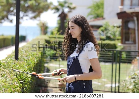 Young woman gardener with curly hair pruning plants bushes in the yard near her house. Teenager with pruner or pruning shears cutting branches at summer garden.