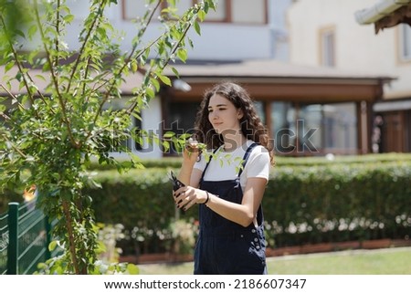 Teen girl gardener with curly hair pruning trees in the yard, working in the garden. Young woman with pruner cutting branches. 