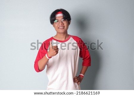 Portrait of attractive Asian man in t-shirt with red and white ribbon on head, showing good job hand gesture with thumbs up. Isolated image on gray background