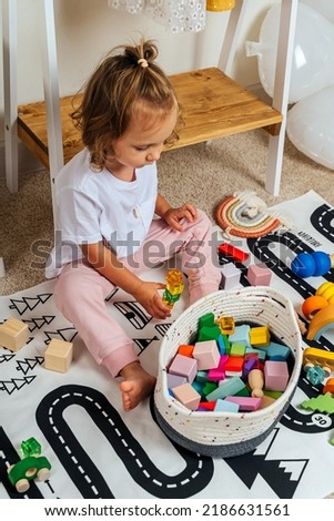 A little girl sitting on floor building tower of block toys on a rug with a picture of a road in playroom. Educational game for baby and toddler in modern nursery. 