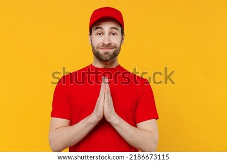 Professional delivery guy employee man wear red cap T-shirt uniform workwear work as dealer courier hold hands folded in prayer gesture, begging isolated on plain yellow background. Service concept. Royalty-Free Stock Photo #2186673115