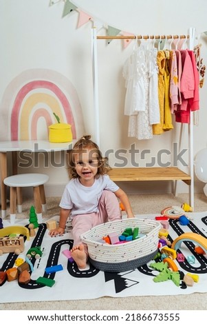 A smilling little girl sitting on floor plays with toy wooden blocks on a rug with a picture of a road in playroom. Educational game for baby and toddler in modern nursery. 