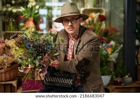 Cute male person in eyeglasses and hat sitting with old typewriter in flower shop. Poet or writer working concept. High quality vertical image