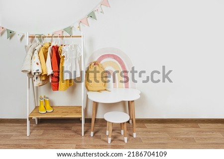 Children's room with Montessori Clothing Rack, white table and rainbow. Dress, jacket and sweaters on hangers in wardrobe. Nursery Storage Ideas. Montessori Toddler Room