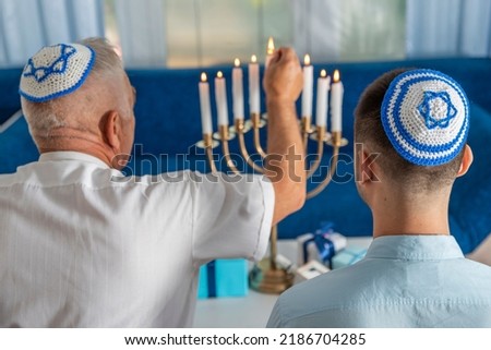 Hanukkah holiday. A senior man and young guy wear kippahs on heads in front of Jewish Hanukkah Menorah, lighting fire of candles. Yarmulke with Star of David. Traditional Hebrew Festival of Lights. Royalty-Free Stock Photo #2186704285