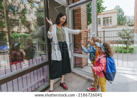 Happy smiling woman teacher greeting elementary school students at first day at school standing near the open door outside. Selective focus. Royalty-Free Stock Photo #2186726041