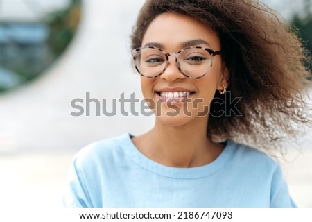 Close-up photo of a beautiful charismatic young African-American woman with curly hair, wearing glasses, with even white teeth, standing outdoors, looking at the camera, smiling friendly