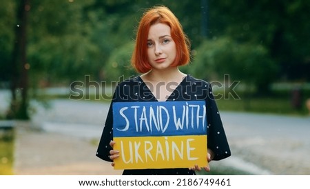 Sad redhead girl hold cardboard with written inscription phrase stand with Ukraine shows patriotic banner painted in ukrainian national blue yellow colors upset anxious young woman ask active