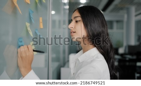 Thoughtful young businesswoman arabian hispanic creative designer come to note board in office read colorful stickers write reminders on sticky papers handwriting to-do list organize corporate goals