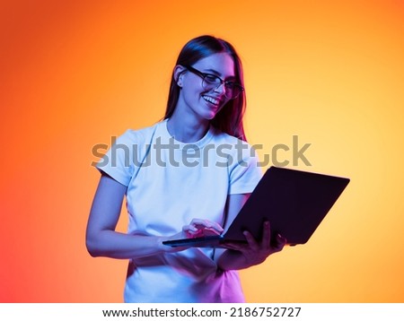 Remote studying. Portrait of young smiling girl, student in white t-shirt with laptop isolated on orange color background in neon light. Concept of beauty, art, fashion, education Royalty-Free Stock Photo #2186752727