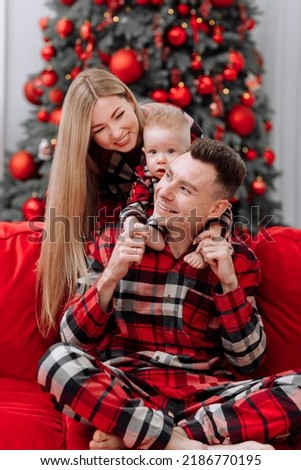 The theme of the family holiday is New year and Christmas. Young European family in the same pyjamas: mom, dad, baby boy are sitting on the red sofa by the festive Christmas tree on Christmas evening.