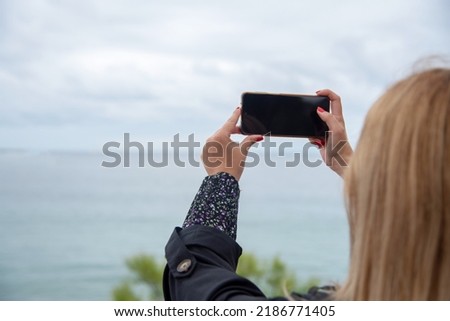 Young blonde woman with red painted nails taking a picture of the landscape with her mobile phone on a cloudy day. Tourism, new technologies, development and lifestyle