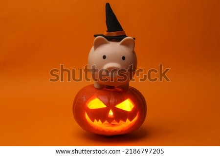 Halloween pumpkin head jack lantern with burning candles and piggy bank with witch hat on orange background. Creative halloween still life