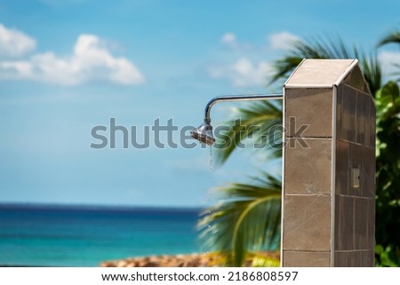 Photo of a metallic shower head in the open air, drops of clear water are falling down from it, you can see the ocean in a blur in the background, and some leaves at the right part of the picture