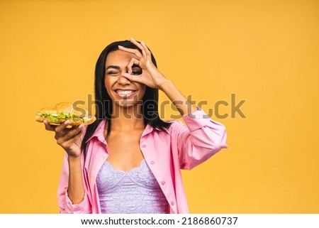 African American black beautiful young woman eating croissant or sandwich isolated over yellow background. Unhealthy junk food diet concept.