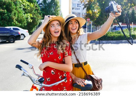 Outdoor portraits of two pretty girls friend taking selfie on vintage camera, enjoy nice day,welling with retro bikes, wearing elegant hats and dresses, looking on camera and making funny faces.