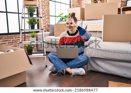 Young hispanic girl sitting on the floor at new home with laptop smiling happy doing ok sign with hand on eye looking through fingers 