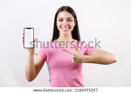 Young beautiful brunette woman with long hair wearing pink t-shirt, pointing at a phone over isolated background. Portrait of female model showing blank screen cellphone. Close up, copy space