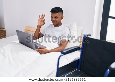 Young hispanic man lying on the bed, using wheelchair doing ok sign with fingers, smiling friendly gesturing excellent symbol 