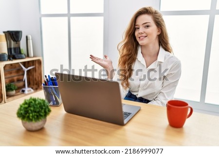 Young caucasian woman working at the office using computer laptop smiling cheerful presenting and pointing with palm of hand looking at the camera. 