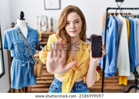 Young caucasian woman at retail shop using smartphone with open hand doing stop sign with serious and confident expression, defense gesture 